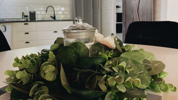 A kitchen with white cabinets, a window with curtains, a sink, and a table with a glass jar and green plants.
