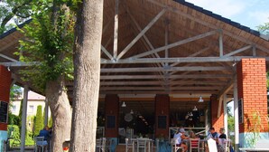 An outdoor cafe with a wooden roof and brick walls, featuring tables and chairs for dining.