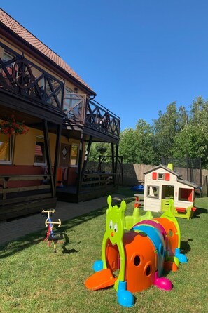 Ein Holzhaus mit Balkon, ein Spielhäuschen und ein grünes Spielzeugfahrzeug auf der Rasenfläche.