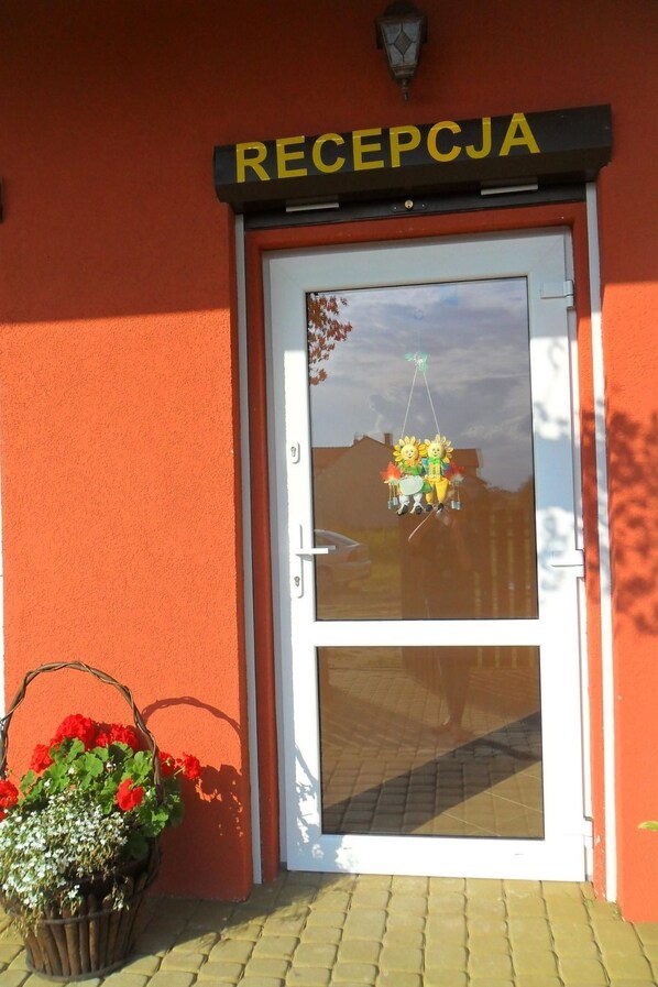 A reception area with a sign reading "RECEPCJA" above a glass door. There is a reflection of a person and a building in the glass, and a basket of flowers is placed outside the door. - Appartement mit einem Wohn-,Ess- und Schlafraum und kleiner Terrasse für vier P by Interhome (Niechorze)