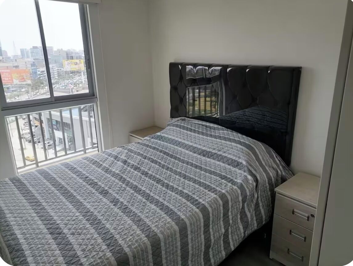 A bedroom with a bed, a nightstand, and a dresser. There is a window with a view of buildings outside.