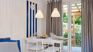 A dining area with a table set for four, white chairs, a clock on the wall, and a view of greenery through a glass door. - Bootshaus mit Schwimmbad by Interhome (Rewal)