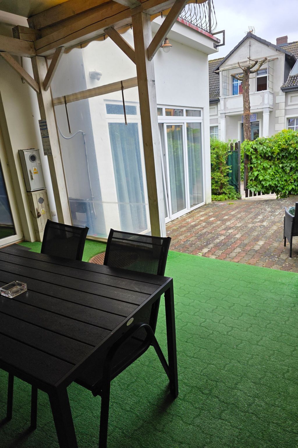 Un patio exterior con una mesa y sillas de madera, suelo de césped artificial verde y una puerta de cristal que da a una casa.