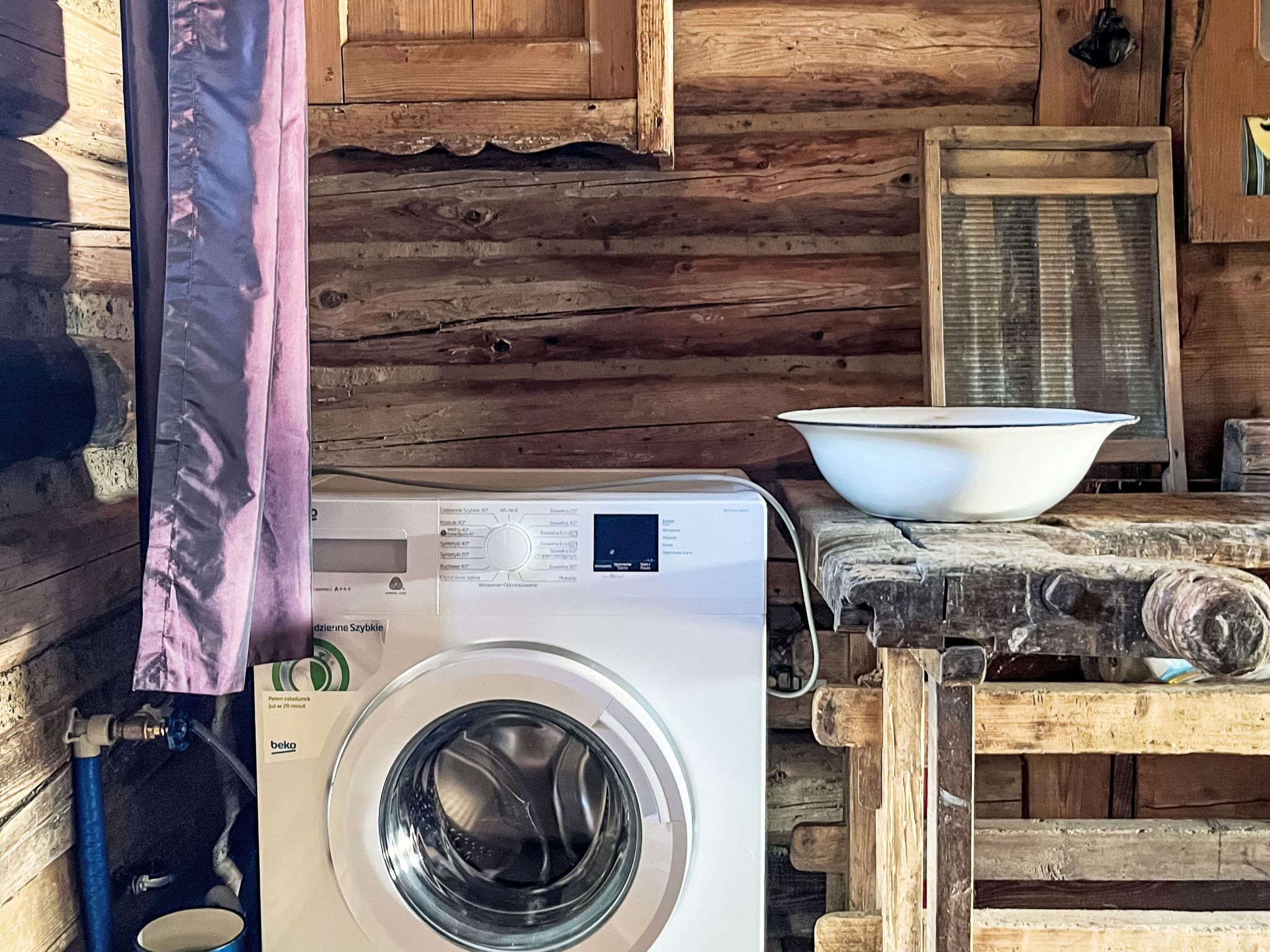 A rustic cabin interior with a washing machine, a wooden table with a bowl, and a window with a screen.