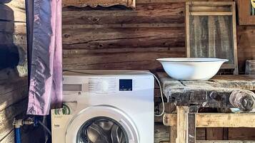 A rustic cabin interior with a washing machine, a wooden table with a bowl, and a window with a screen.