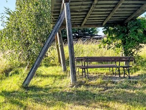 A wooden bench under a shelter with a metal frame, surrounded by grass and trees.