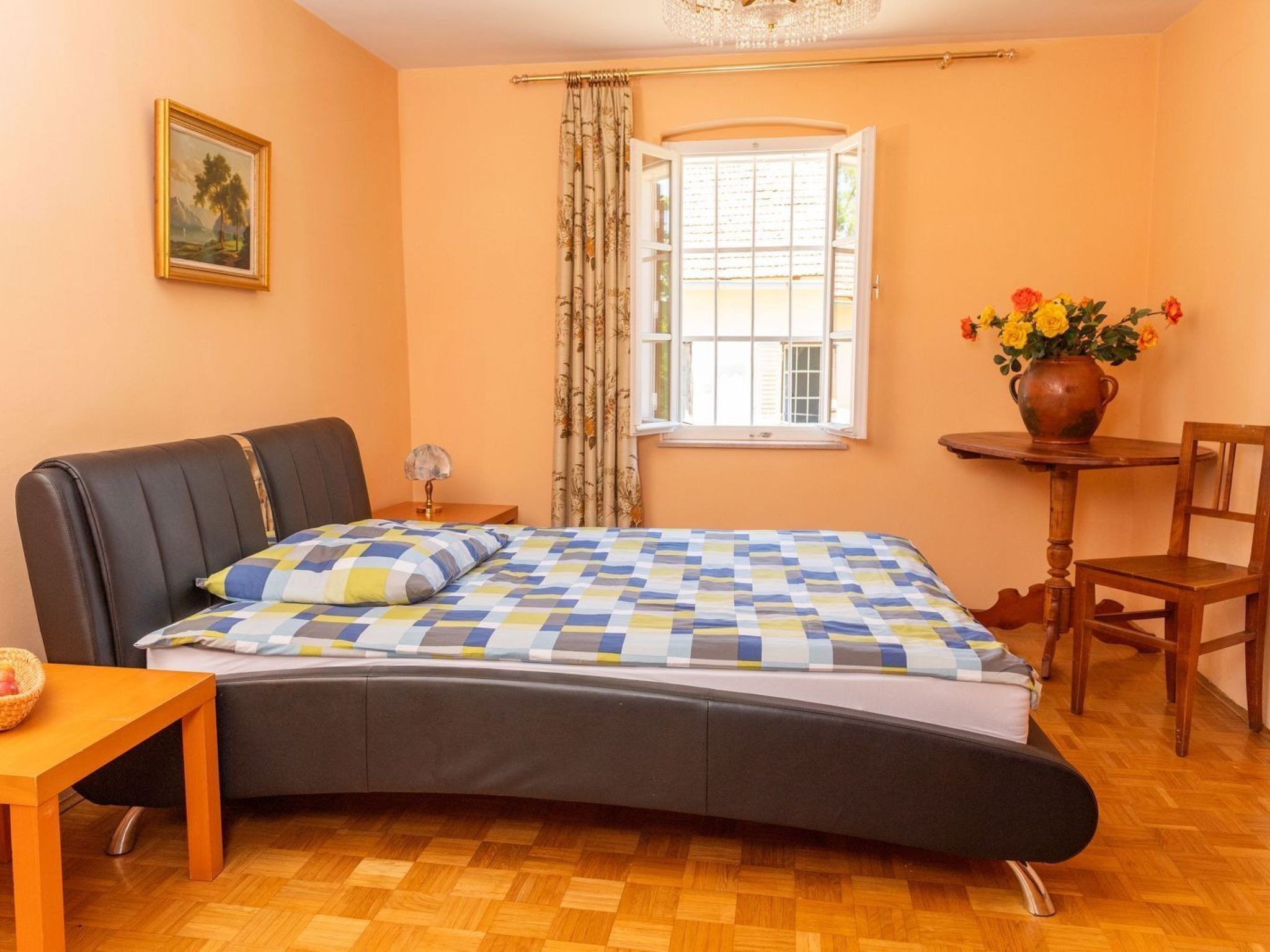 A bedroom with a bed, a nightstand, a chair, a table, a window with curtains, and a vase with flowers.