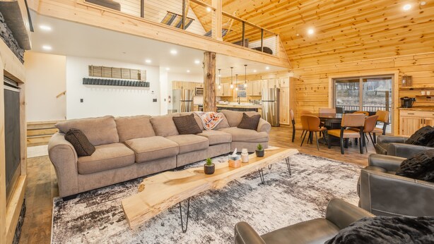 A spacious living area with a large sectional sofa, a wooden coffee table, and a high ceiling with exposed wooden beams. - Brand New Luxury 7 BR Cabin w Hot Tub, Game Room (McHenry)