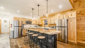 A modern kitchen with wooden cabinets, stainless steel appliances, a central island with stools, and pendant lighting. - Brand New Luxury 7 BR Cabin w Hot Tub, Game Room (McHenry)
