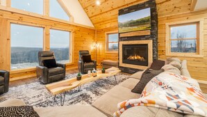 A log cabin interior with a fireplace, wooden walls, and large windows offering a view of the outdoors. - Brand New Luxury 7 BR Cabin w Hot Tub, Game Room (McHenry)