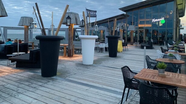 An outdoor deck with wooden flooring, black and white outdoor seating, and a modern building with glass windows in the background. - in Dirkshorn by Interhome (Dirkshorn)