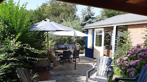 Un patio al aire libre con una mesa y sillas, una zona de estar cubierta y un pequeño edificio con fachada azul.