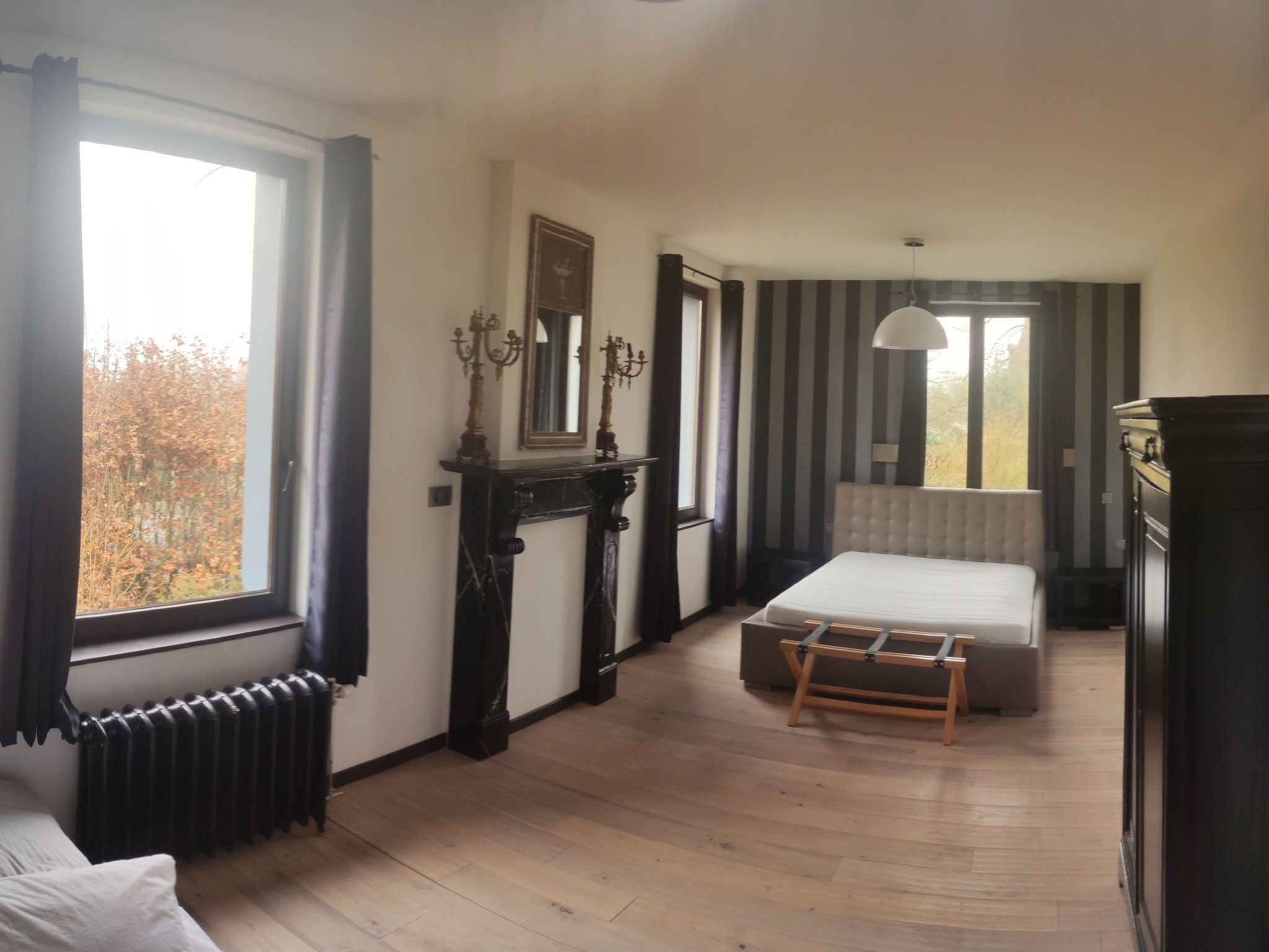 A bedroom with a large window, a radiator, a bed, a bedside table, and a mirror.