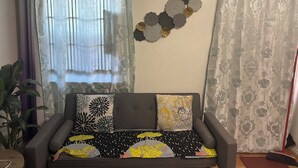 Een woonkamer met een bank, decoratieve kussens, een wandairconditioner en gordijnen met een patroon.