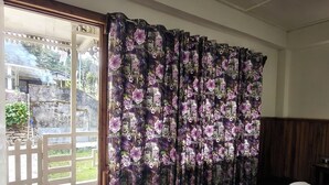 Una habitación con un gran ventanal cubierto por cortinas florales, piso de madera y una cama con colcha estampada.