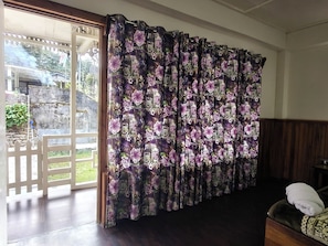 Una habitación con un gran ventanal cubierto por cortinas florales, piso de madera y una cama con colcha estampada.