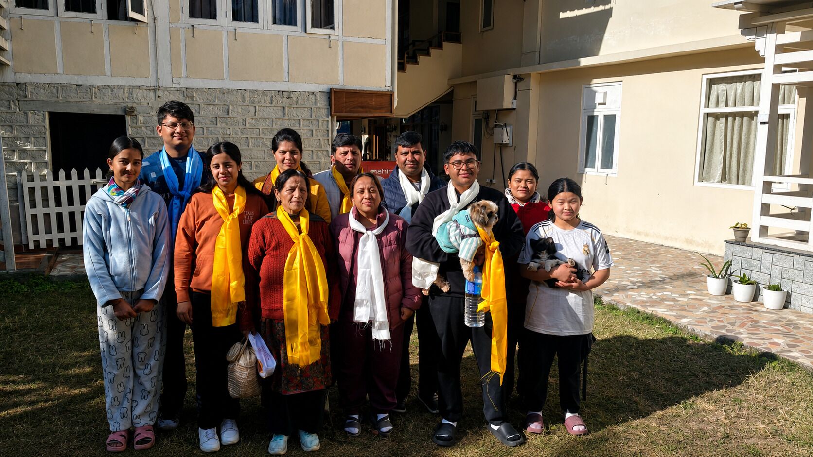A group of people standing in front of a building with a white picket fence, a tiled entrance, and a small dog being held by one individual.