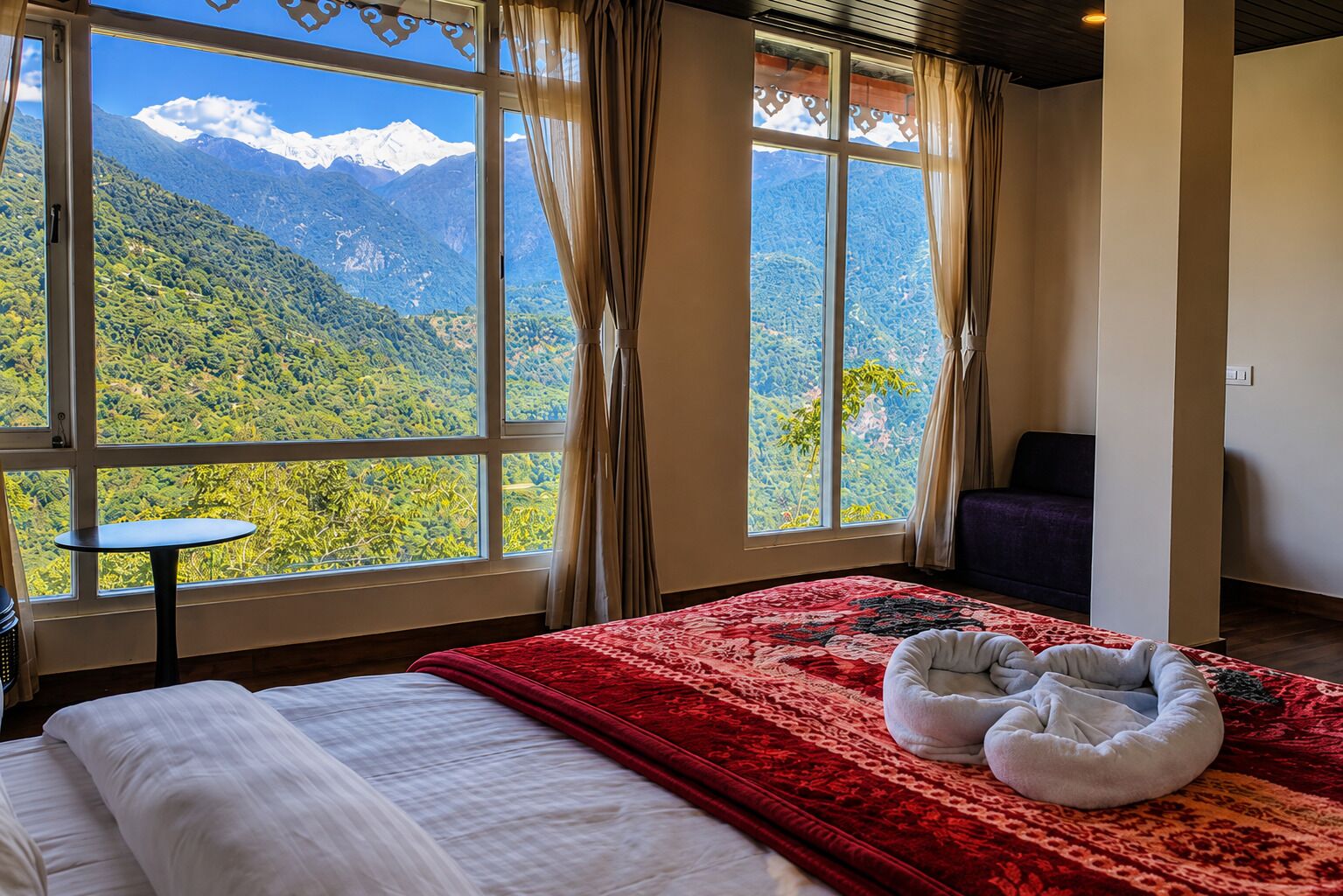 A bedroom with a large window offering a mountain view, a bed with a red blanket, a small table, and a pet bed.