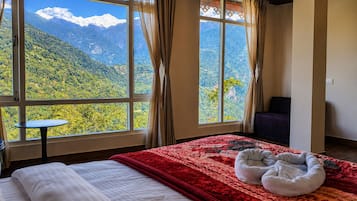 A bedroom with a large window offering a mountain view, a bed with a red blanket, a small table, and a pet bed.
