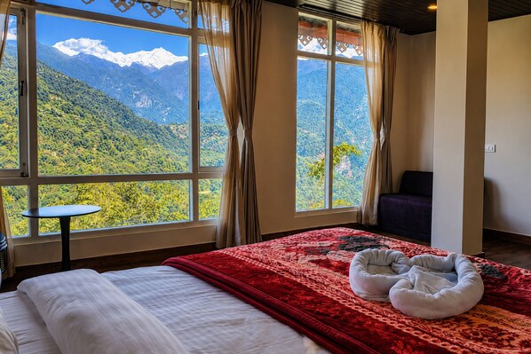 Un dormitorio con un ventanal grande que ofrece vistas a la montaña, una cama con manta roja, una mesita y una cama para mascotas.