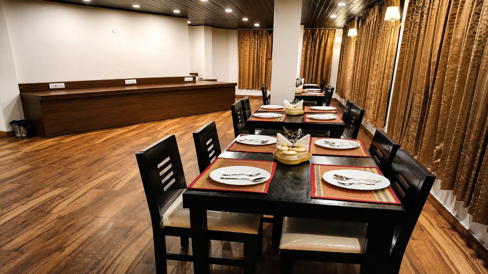 Un comedor con una larga mesa dispuesta para varios comensales, suelo de madera y un mostrador de recepción al fondo.