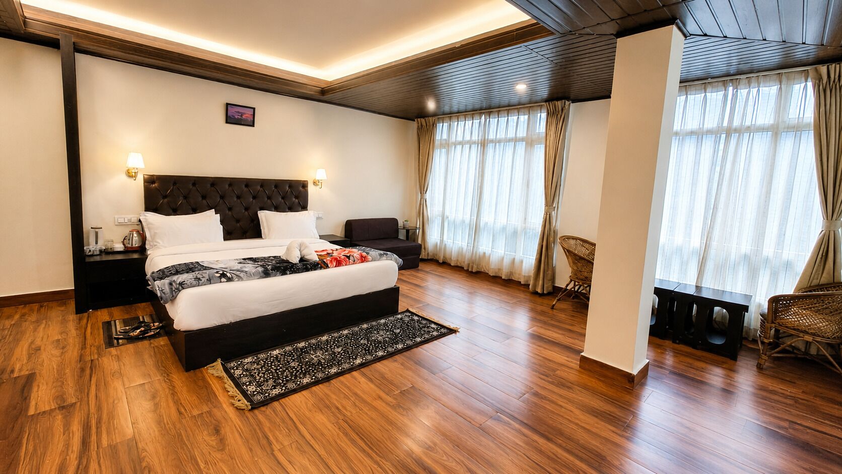 A modern hotel room with a large bed, wooden flooring, and a view of the outdoors through sheer curtains.