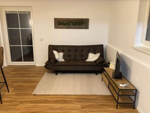 Ein Wohnzimmer mit einem dunkelbraunen Sofa, zwei weißen, flauschigen Kissen, einem weißen Teppich, einem Holztisch und einem Fernseher auf einem Schrank.