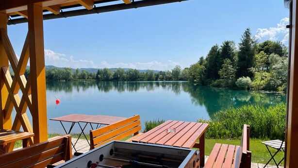 An outdoor seating area with a foosball table, wooden benches, and a view of a calm lake surrounded by greenery. - Apartment Ahorn by Interhome (Feistritz im Rosental)