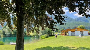 A lakeside lodge with a wooden cabin, a grassy lawn, and a clear view of the lake and mountains in the background. - Apartment Esche by Interhome (Feistritz im Rosental)