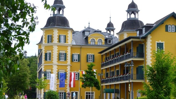 A yellow building with multiple balconies and a central tower. Flags are displayed outside. - Apartment Esche by Interhome (Feistritz im Rosental)