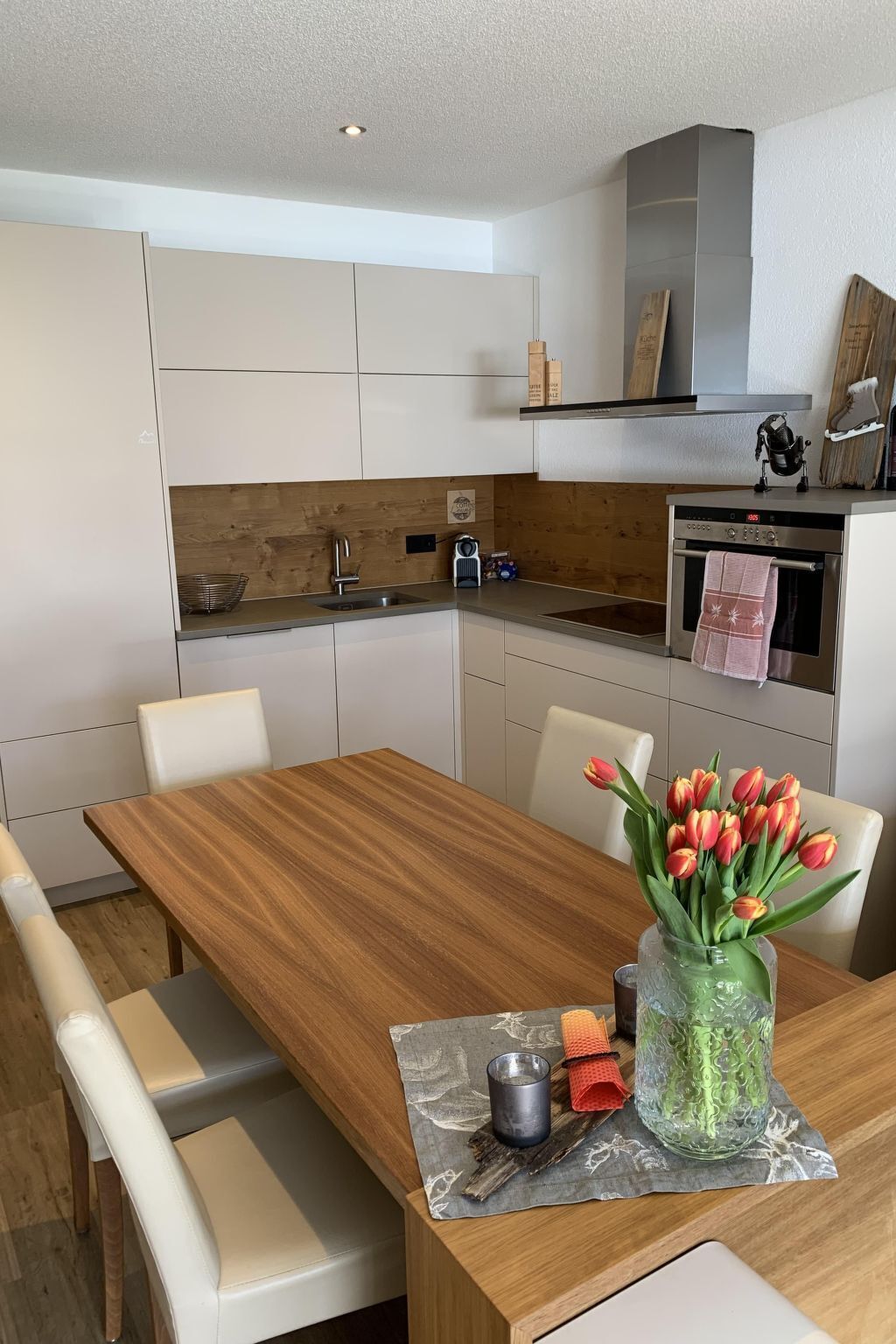 A modern kitchen with white cabinets, a wooden countertop, and a built-in oven. There is a dining table with chairs and a vase of tulips.
