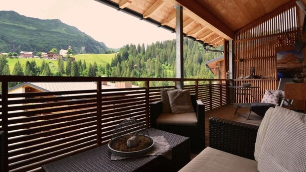 Un balcon donnant sur une vallée verdoyante et des montagnes, avec des balustrades en bois, des meubles en osier et une petite table agrémentée d’un objet décoratif.