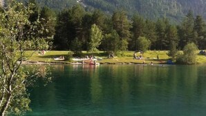 Un lago con montañas al fondo, una zona verde con gente y una pequeña embarcación.