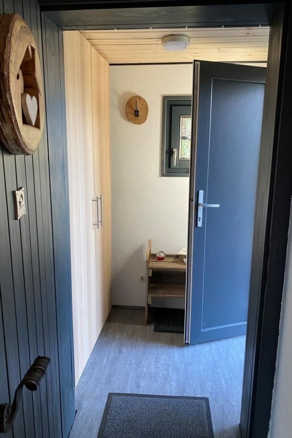 A narrow hallway with a wooden door on the right, a small wooden shelf with a red object, and a wall-mounted clock.