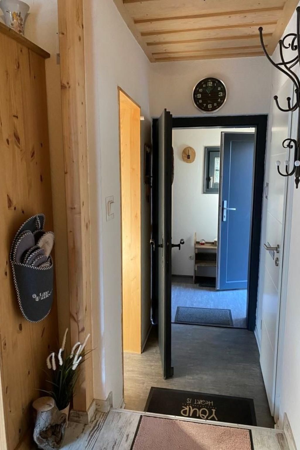 A hallway with a wooden door leading to a room, a clock on the wall, and a doormat with text.