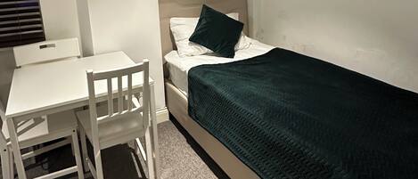 A small, neatly arranged room with a single bed, a bedside table, a chair, and a desk.