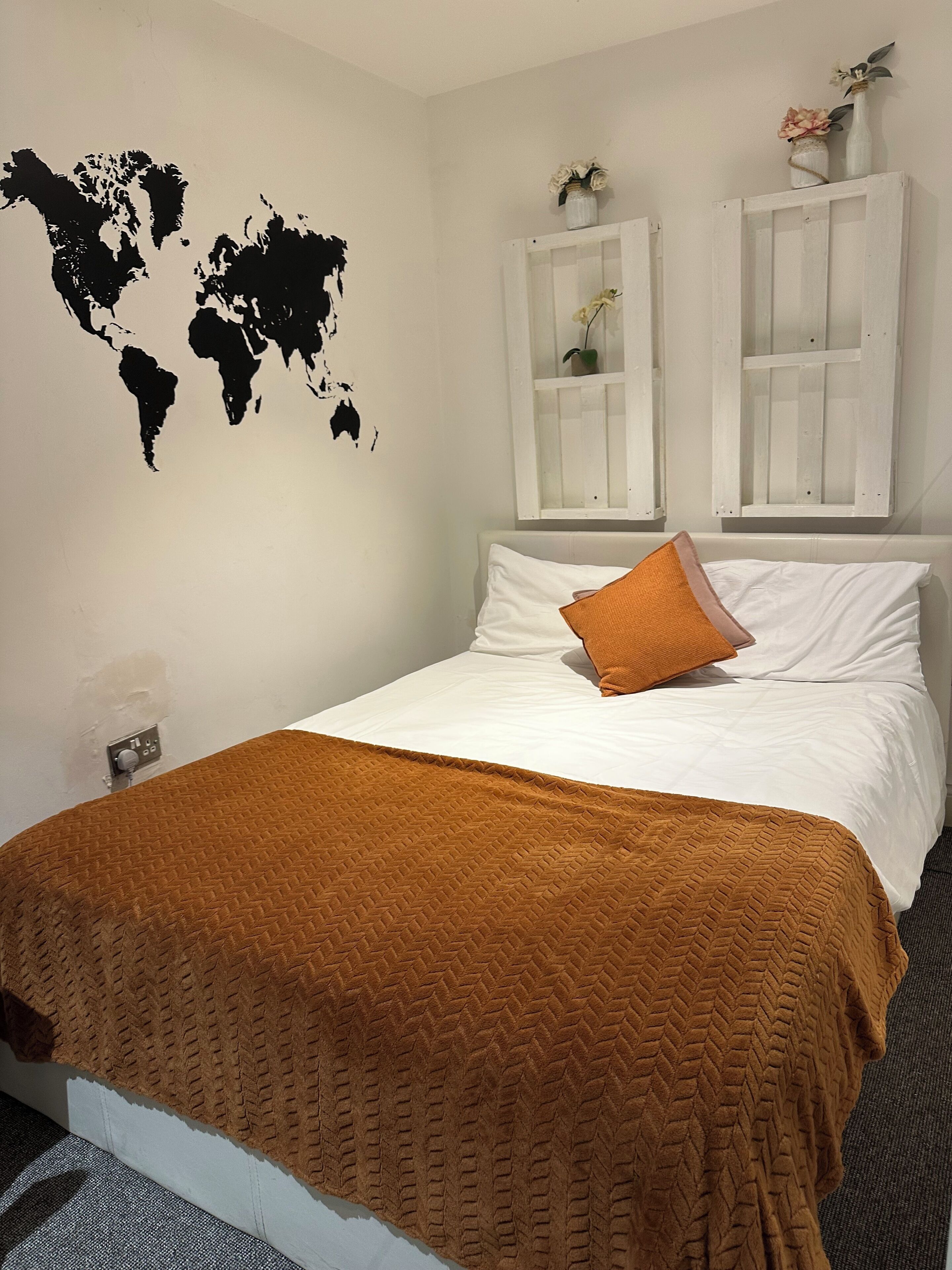 A neatly made bed with a brown blanket, white pillows, and a single orange pillow. There are two white cabinets with floral decorations on top. A world map is painted on the wall.