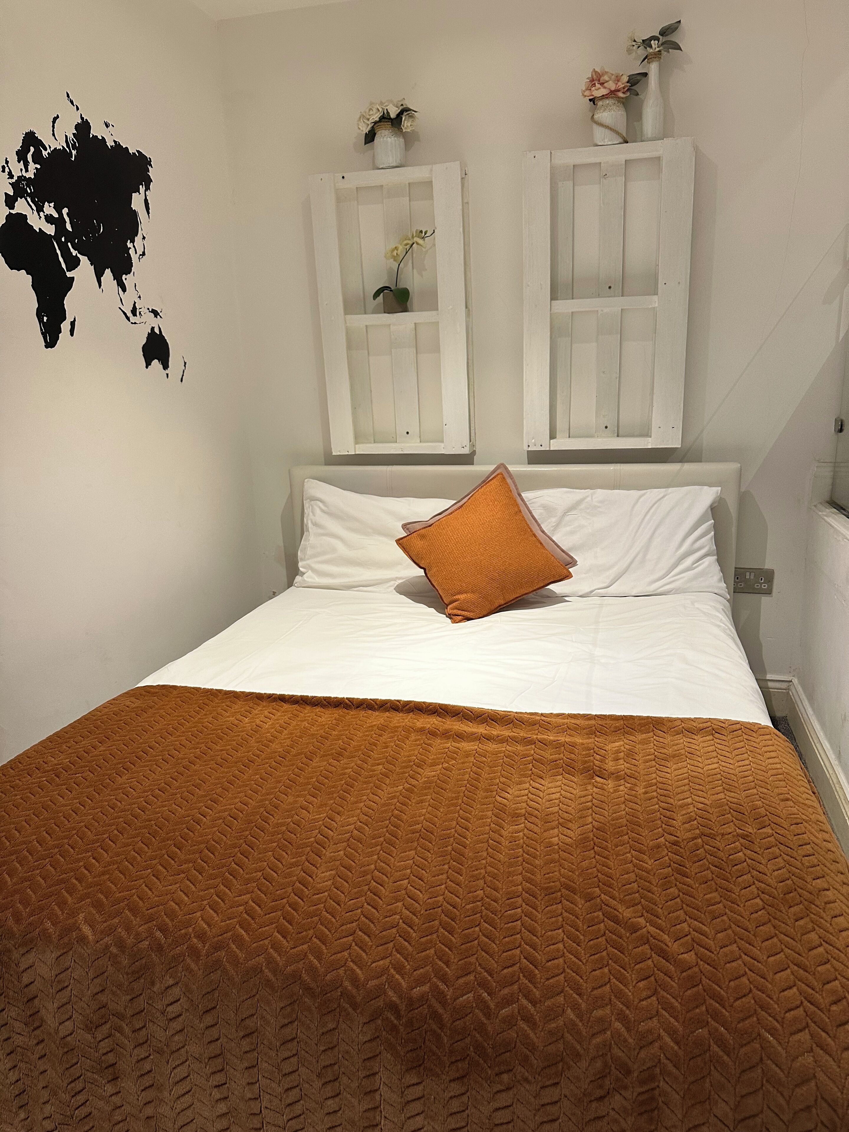 Ein ordentlich bezogenes Bett mit einer braunen Decke, weißen Kissen und einem einzelnen orangen Kissen. An der Wand stehen zwei weiße Schränke mit Glastüren, in jedem befindet sich eine Pflanze.