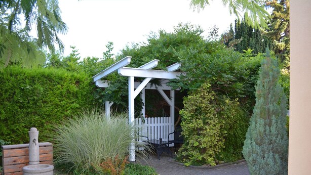 Un jardin agrémenté d’une pergola blanche, d’un bac à fleurs en pierre et d’une haie.