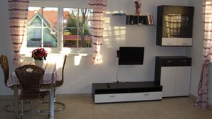 Un espace de vie compact comprenant un coin repas, un téléviseur fixé au mur, un meuble intégré et une fenêtre avec des rideaux.