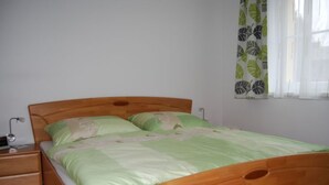 Un lit simple avec un cadre en bois, une couvre-lit verte et des oreillers assortis. Il y a une table de chevet avec une lampe et un tiroir. On aperçoit une fenêtre avec un rideau à motifs de feuilles.