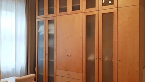 Ein Holzkleiderschrank mit mehreren Türen und Schubladen, ein kleiner Tisch mit Stühlen sowie ein Fenster mit durchsichtigen Vorhängen.