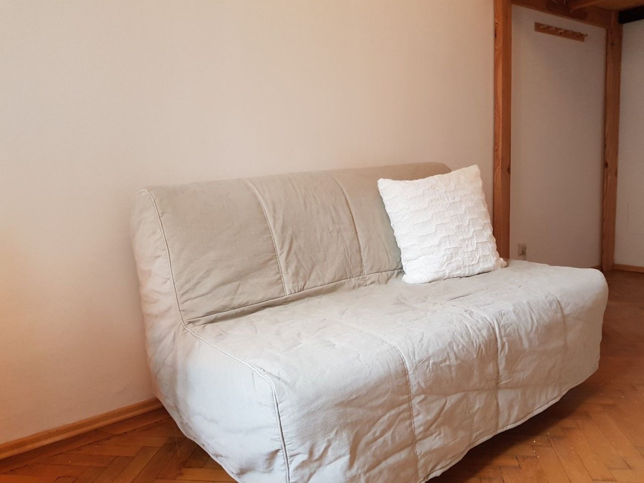 Un canapé de couleur claire, doté d’un revêtement matelassé et d’un seul coussin blanc, dans une pièce au parquet en bois et aux murs unis.