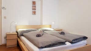 Ein ordentlich bezogenes Bett mit zwei Kissen und einer Decke, ein Nachttisch mit Lampe sowie eine kleine Kommode.