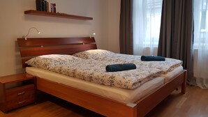 Una cama doble con cabecera de madera, dos almohadas, una mesita de noche, una estantería con libros y una ventana con cortinas.
