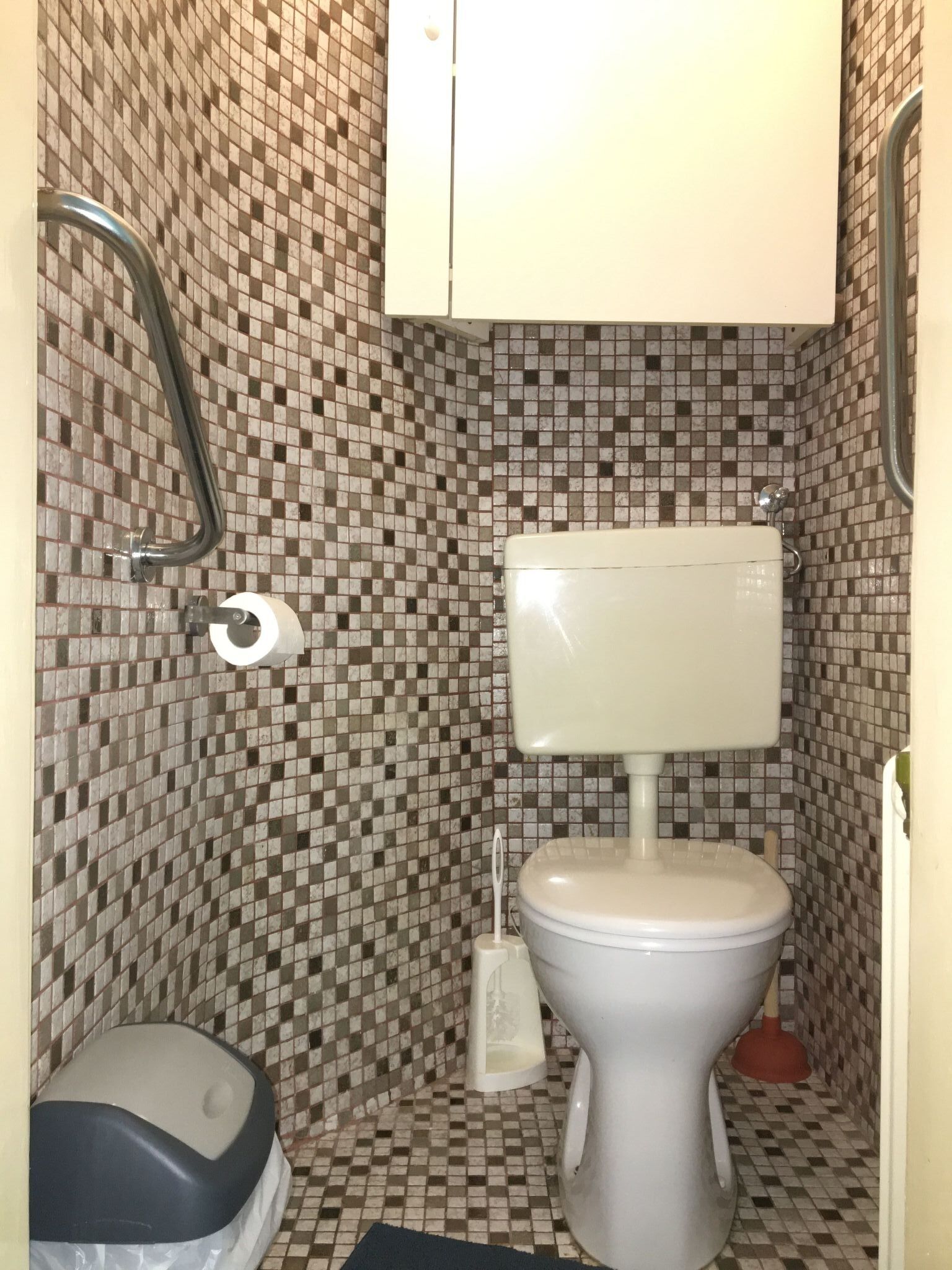 A bathroom with a tiled floor, a toilet, a hand dryer, and a plunger.