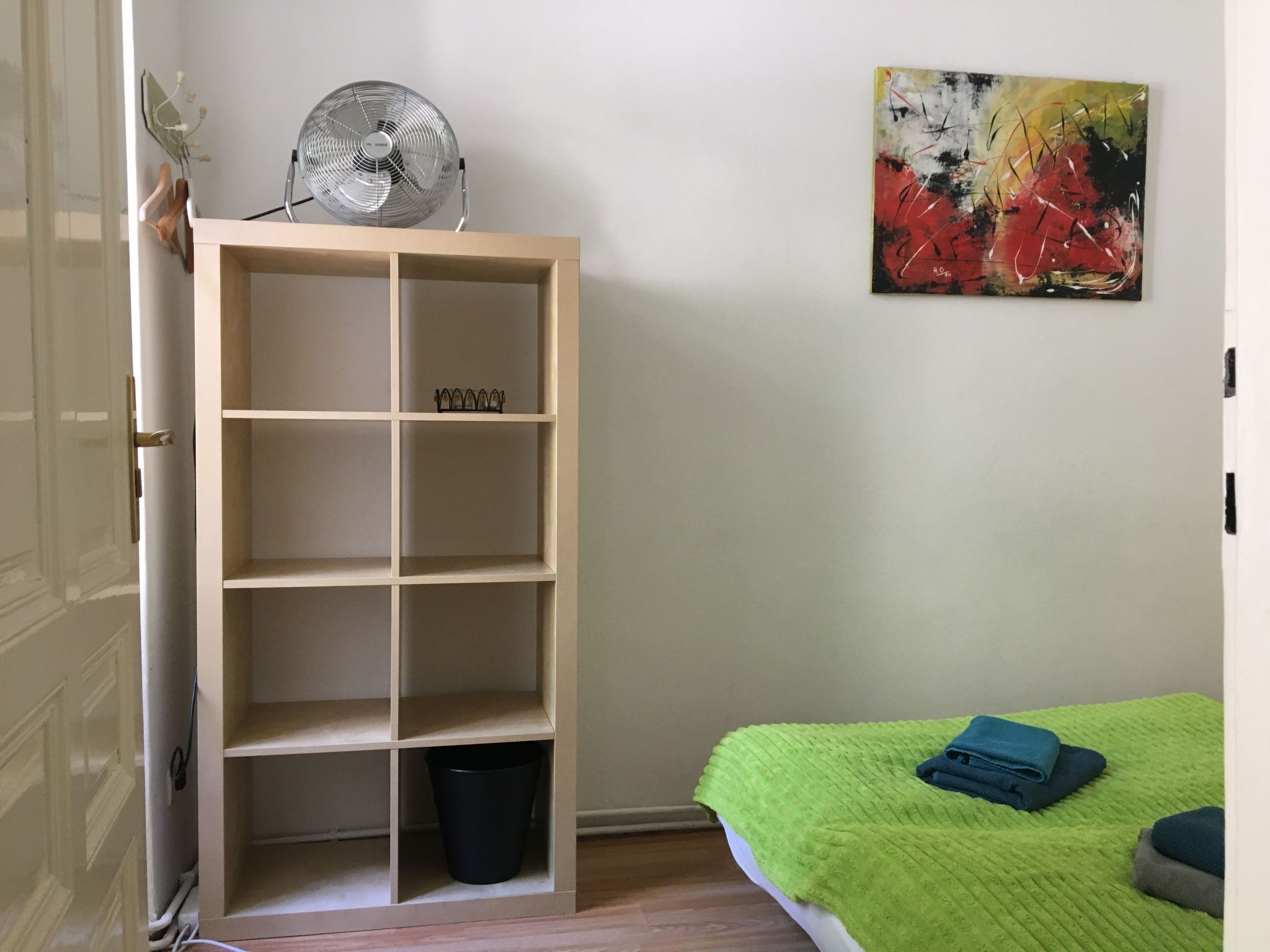 A room with a wooden shelving unit, a fan, a black trash bin, a green blanket, and a colorful abstract painting on the wall.