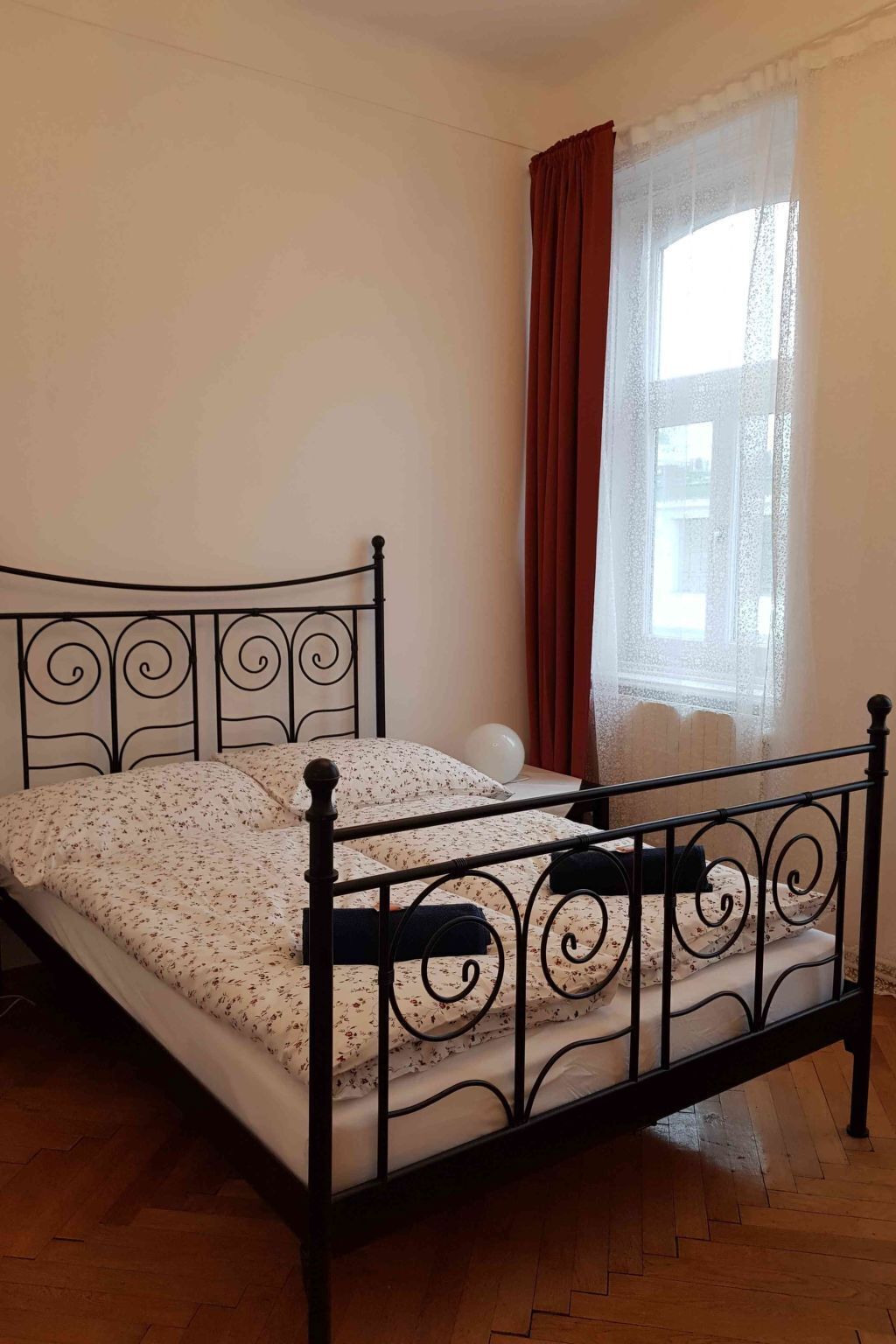 A single bed with a metal frame and a floral bedspread in a room with wooden flooring and a window with red curtains.