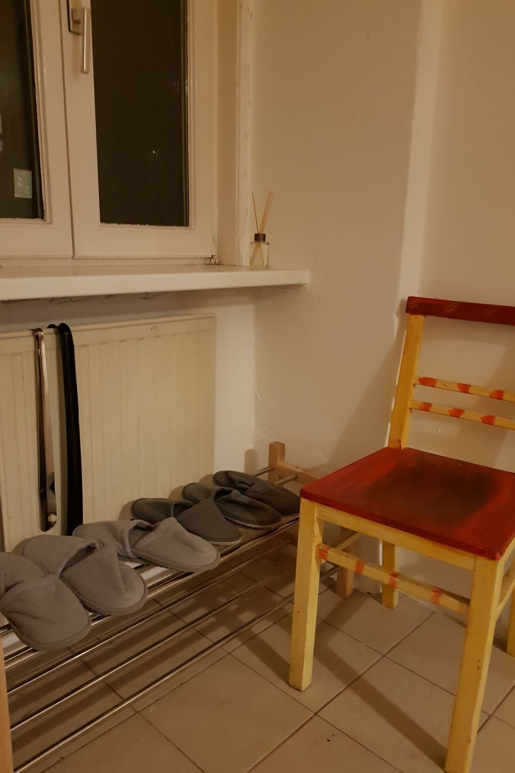 Ein Zimmer mit Heizkörper, Schuhablage und Hausschuhen sowie ein Holzhocker mit rotem Kissen.