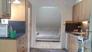 Cocina compacta con armarios de madera, microondas y lavamanos. Un sofá se encuentra en una zona semi-privada con vistas al exterior.